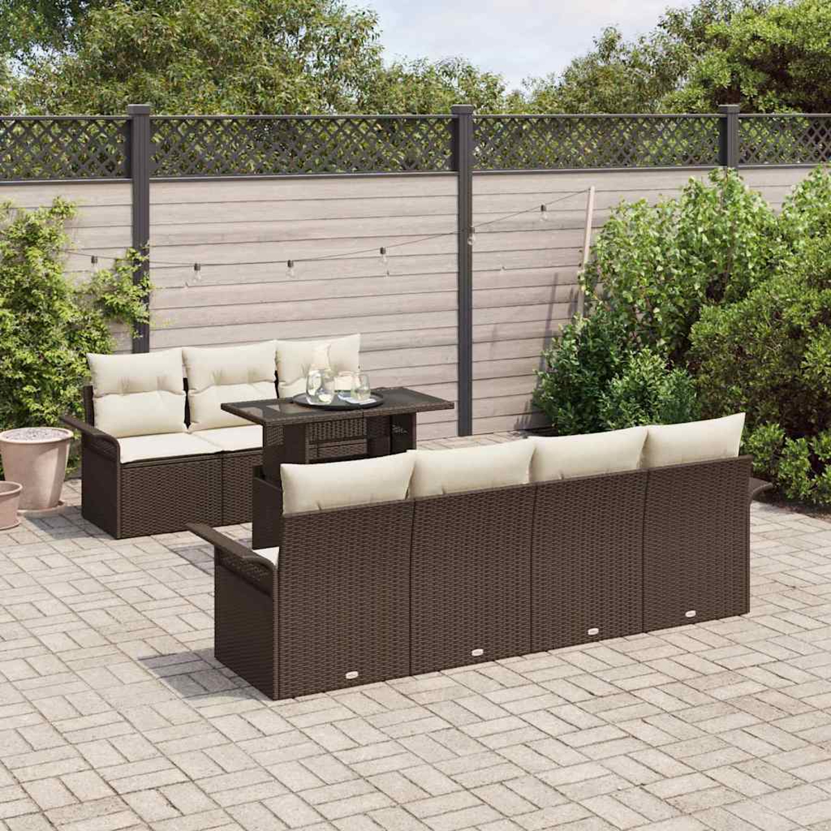 Garten-Terrassenset mit braunen Korbsesseln, beigen Kissen und einem Tisch mit Glasplatte. Vor einem Holzzaun und Grün.