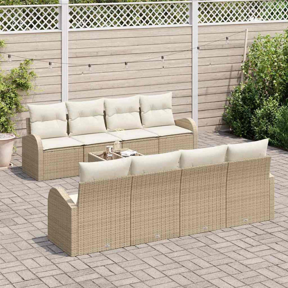 Rattan-Sofa-Set im Freien mit cremefarbenen Kissen, Glastisch und Lichterketten. Auf einer Terrasse mit Holzzaun und Grünanlage.