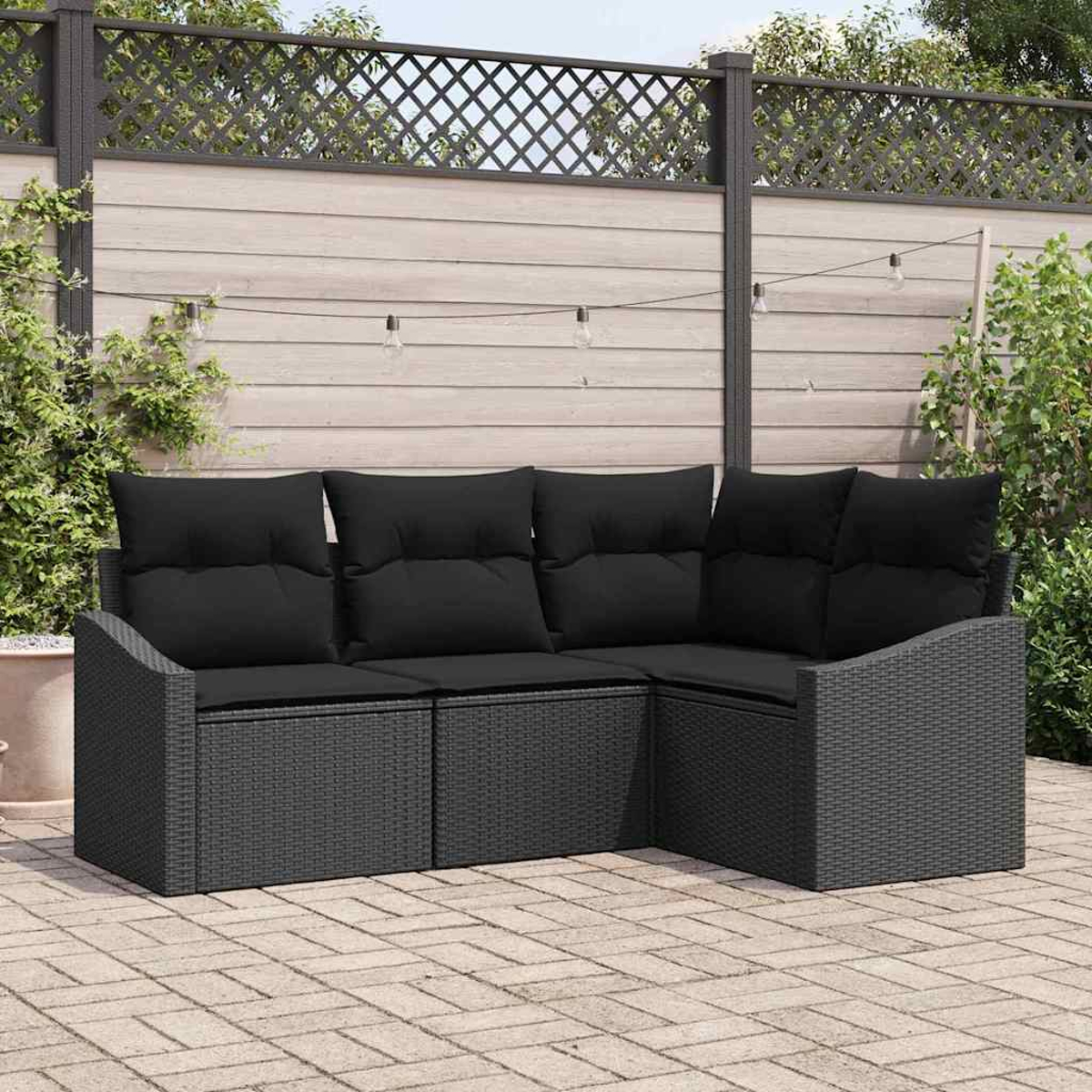 Graues Ecksofa aus Korbgeflecht mit schwarzen Kissen steht auf einer Ziegelterrasse. Im Hintergrund ein Holzzaun und Grün.