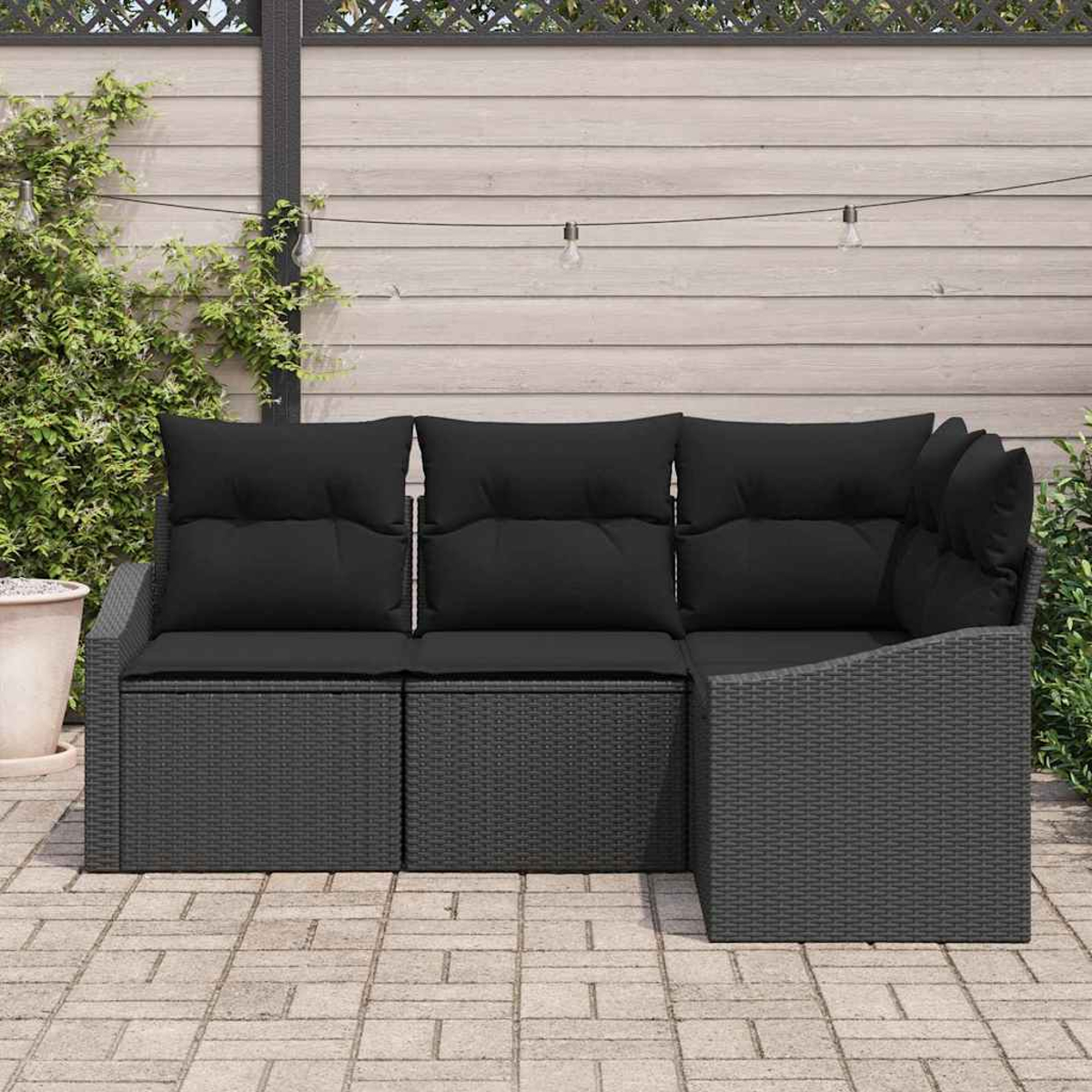 Ein Ecksofa mit schwarzen Kissen steht auf einer Terrasse. Im Hintergrund sind ein Holzzaun, Grün und Lichterketten zu sehen.
