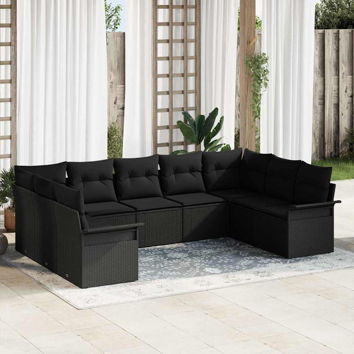 Schwarzes Ecksofa aus Korbgeflecht mit Kissen auf einer Terrasse. Weiße Vorhänge und ein Holzzaun im Hintergrund. Ein Teppich liegt darunter.