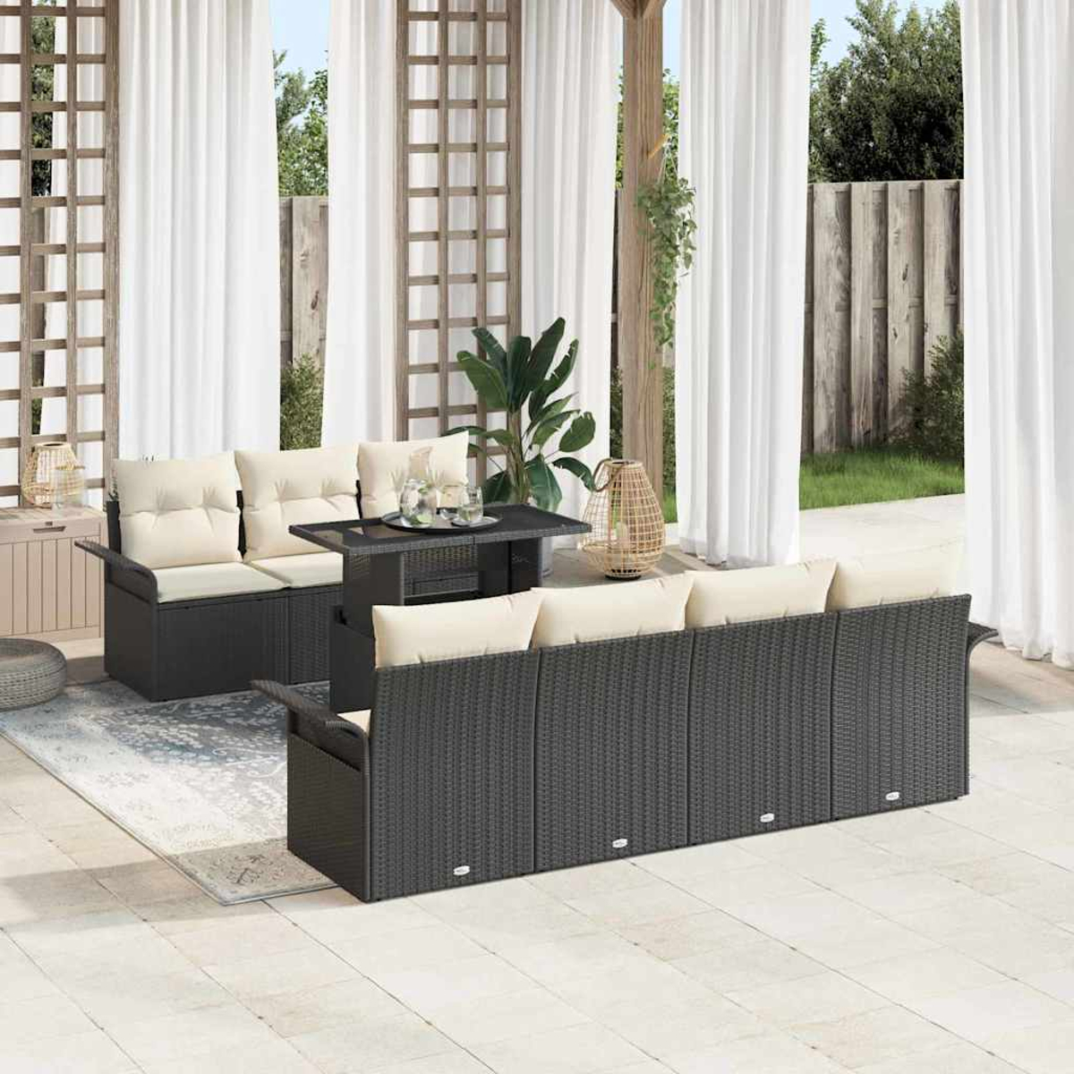 Set patio esterno con mobili in vimini neri, cuscini color crema e un tavolo. Tende bianche, un tappeto e piante verdi completano la scena.