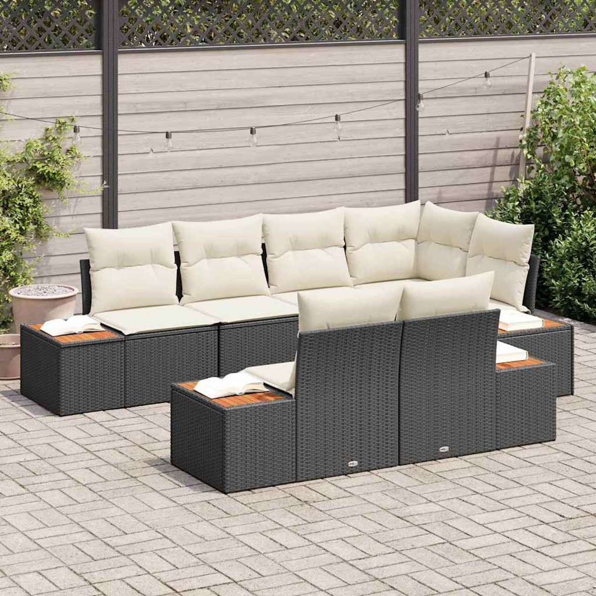 Outdoor-Sitzgruppe. Dunkelgraues Korbmöbel mit cremefarbenen Kissen. Holztische. Gepflasterte Terrasse mit Holzzaun und Grün im Hintergrund.