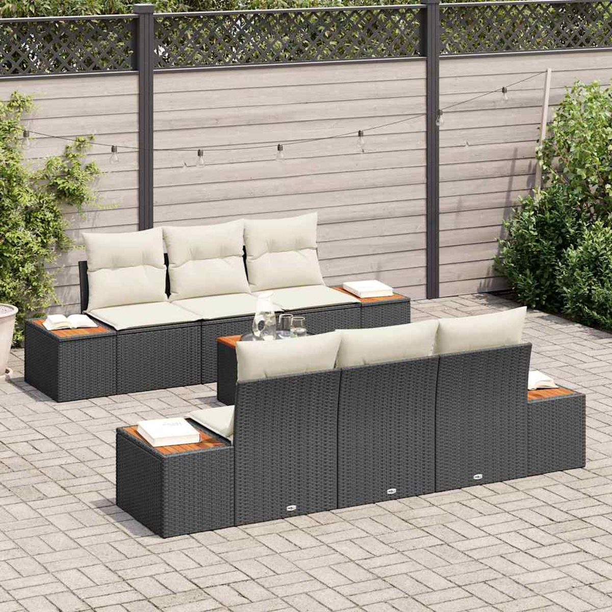 Garten-Lounge-Set mit grauen Korbsesseln, cremefarbenen Kissen und Holztischen. Hintergrund: Holzzaun, Pflanzen und Lichterketten.
