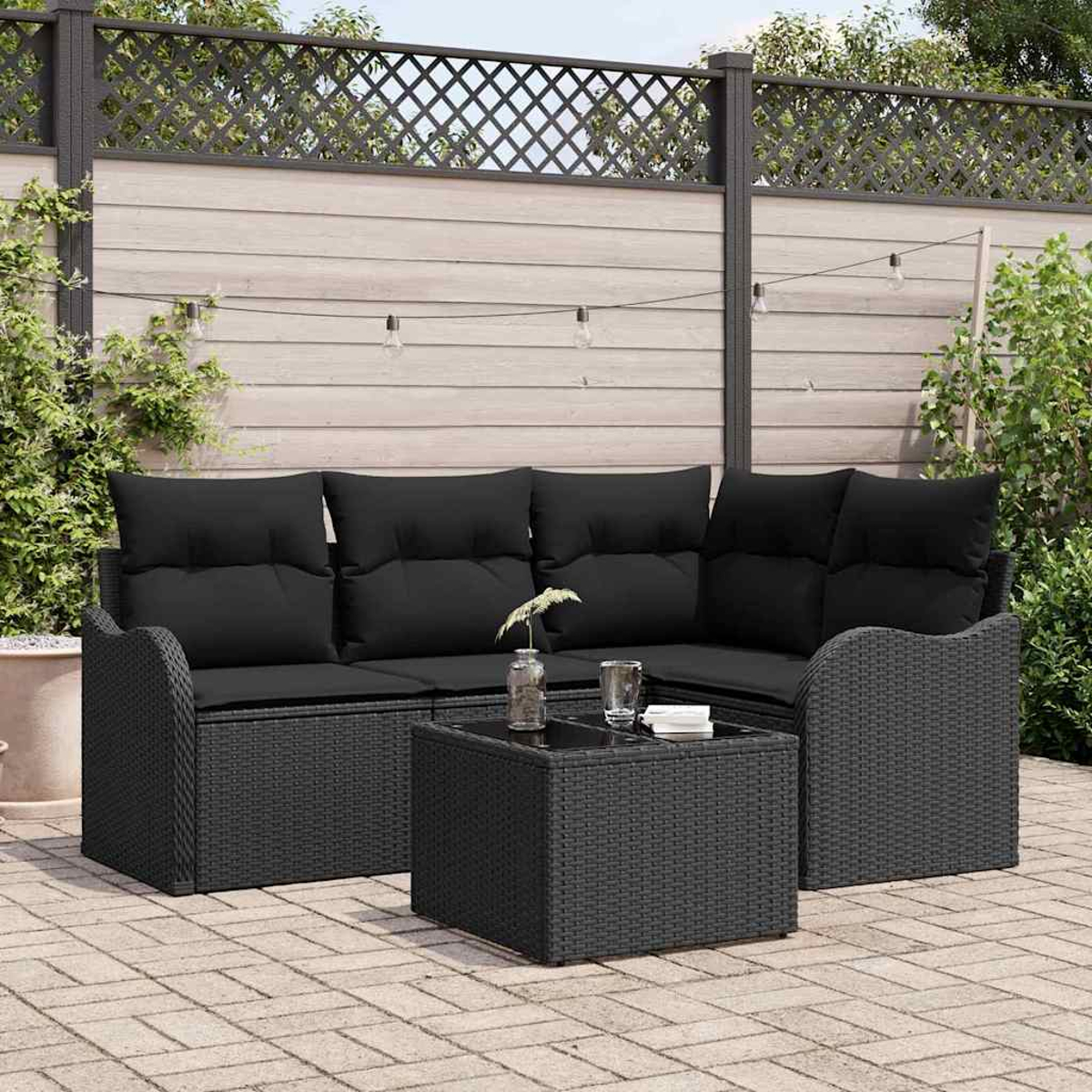Ein schwarzes Outdoor-Ecksofa und -Tisch aus Korbgeflecht auf einer Backstein-Terrasse mit Holzzaun und Grün im Hintergrund.