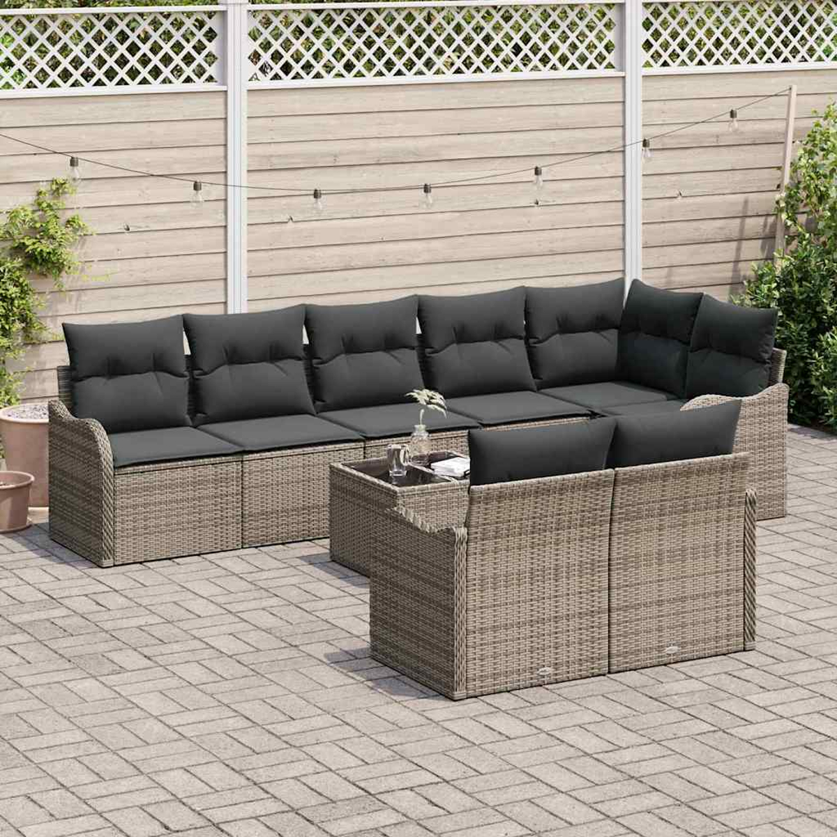 Ein graues Outdoor-Ecksofa aus Korbgeflecht mit schwarzen Kissen und einem kleinen Tisch auf einer Ziegelterrasse mit Holzzaun im Hintergrund.