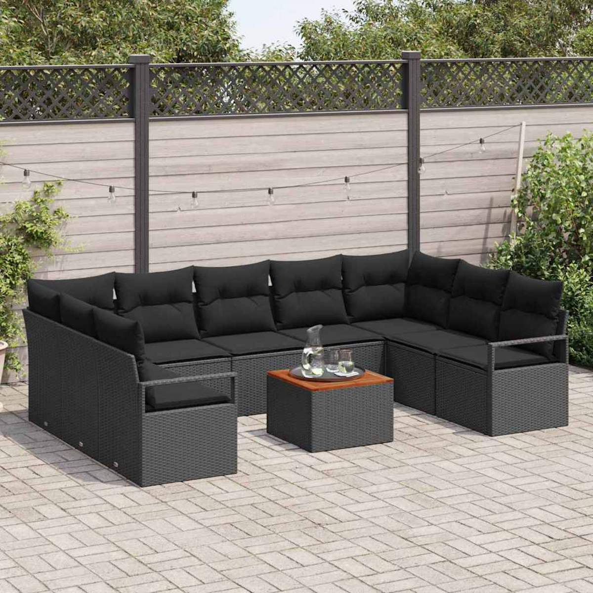 Ein Outdoor-Sitzgarnitur mit schwarzen Kissen. Ein kleiner Tisch mit Holzplatte steht in der Mitte. Im Hintergrund ein Zaun und einige Pflanzen.