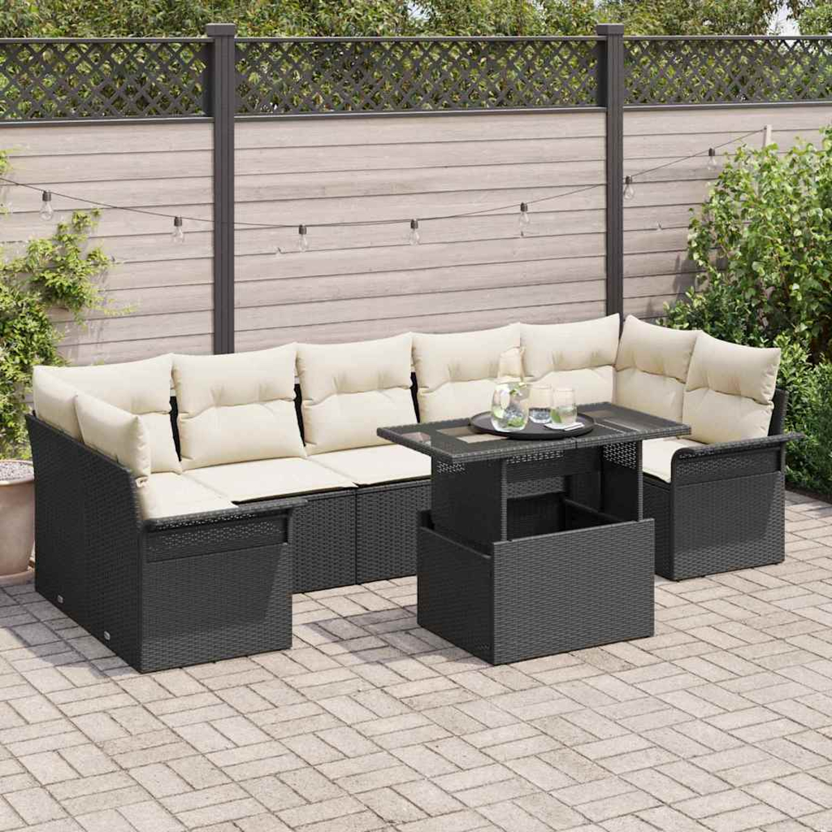 Schwarzes Outdoor-Korbmöbel mit cremefarbenen Kissen, Glastisch und Getränken. Auf einer Backstein-Terrasse mit Zaun und Grün im Hintergrund.