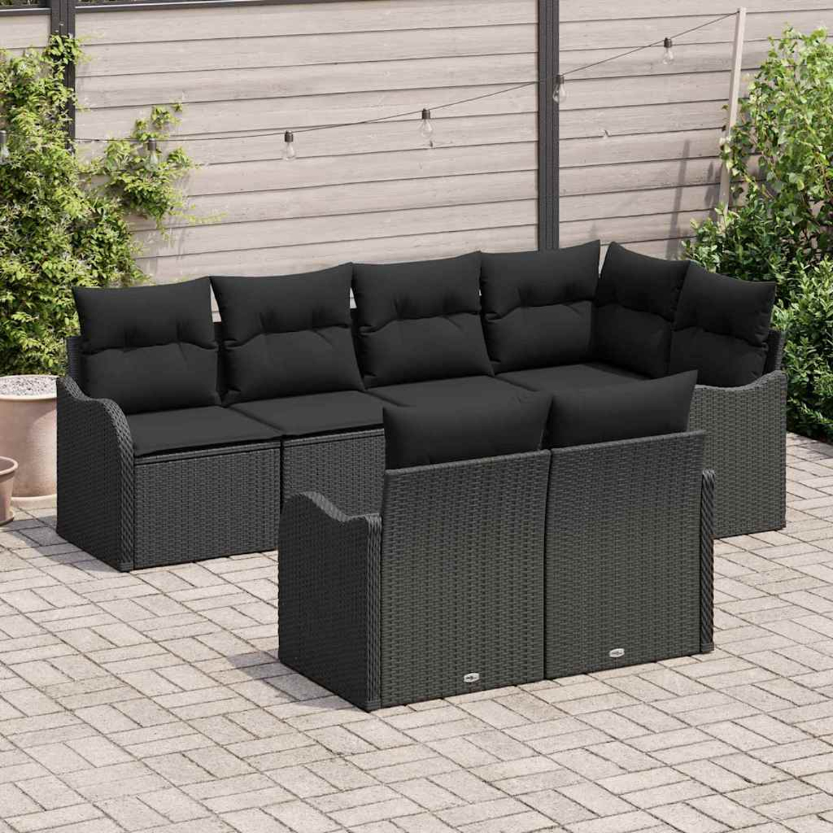Outdoor-Ecksofa mit schwarzen Kissen. Grauer gewebter Rahmen. Terrassenpflaster im Vordergrund. Holzzaun und Pflanzen im Hintergrund.