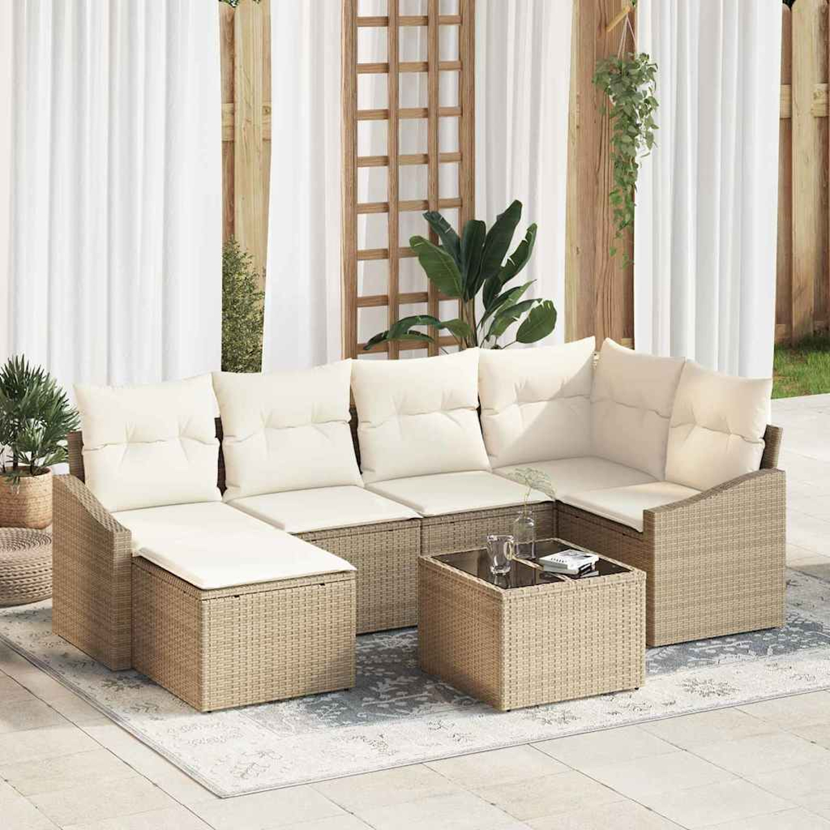 Outdoor-Rattansofa mit cremefarbenen Kissen, Tisch mit Glasplatte, Teppich und Pflanzen. Dahinter weiße Vorhänge, Holzgitter und Zaun.