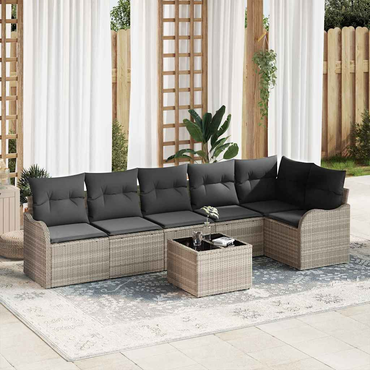 Outdoor-Wicker-Sofa mit grauen Kissen und einem kleinen Tisch auf einem gemusterten Teppich, in der Nähe weißer Vorhänge.