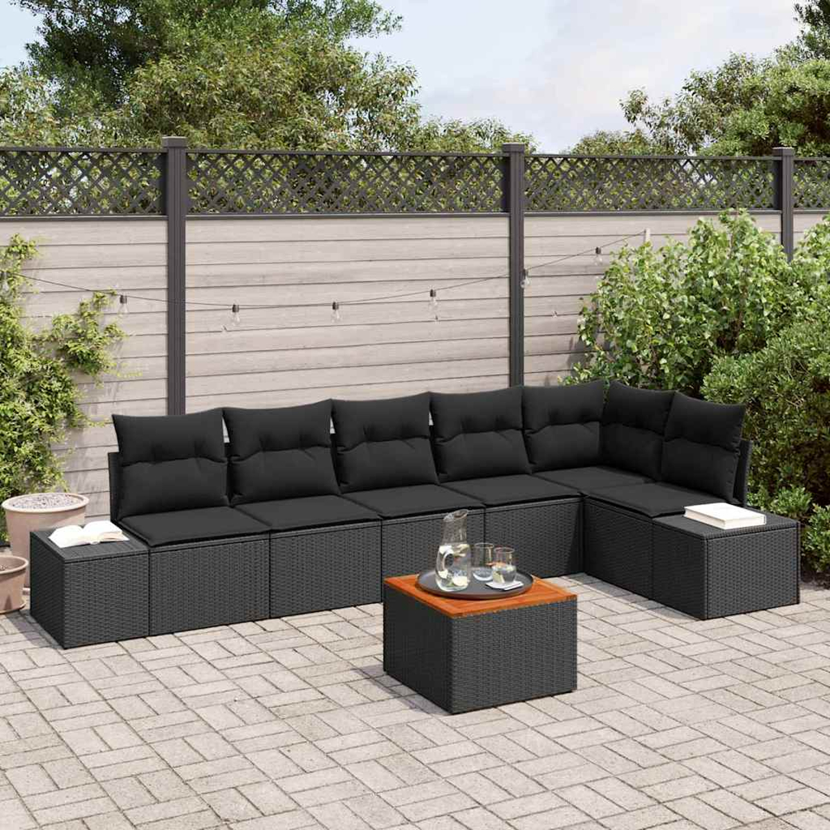 Outdoor-Ecksofa mit schwarzen Kissen, einem kleinen Tisch und einem Krug mit Gläsern auf einer Terrasse. Hintergrund: Zaun und Grün.