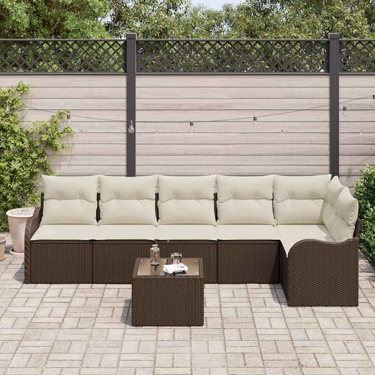 Outdoor-Rattan-Ecksofa mit cremefarbenen Kissen und einem kleinen Tisch. Im Hintergrund befinden sich ein Zaun und Grün.