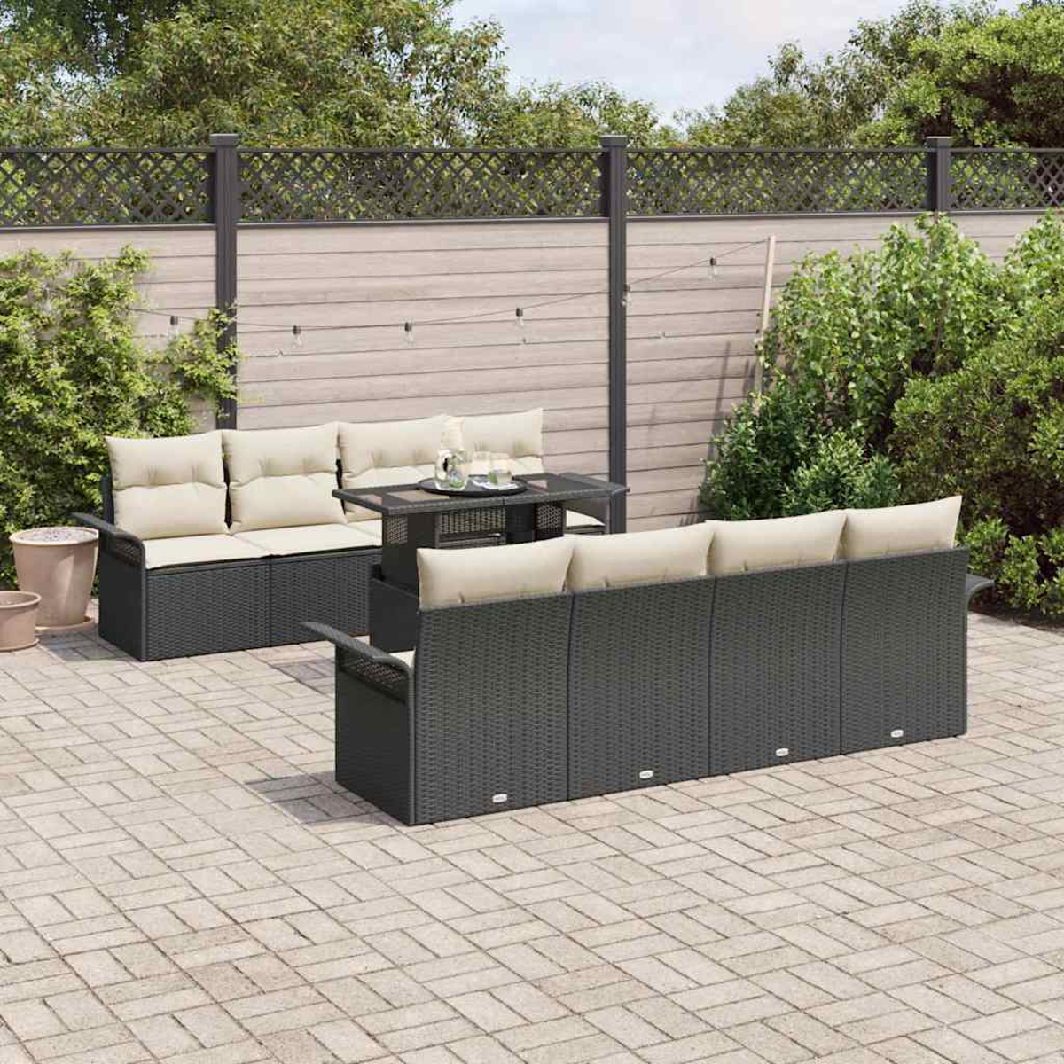 Terrassenset mit dunklen Korbmöbeln, beigen Kissen, kleinem Tisch und gepflasterter Terrasse, mit Zaun und Grün.