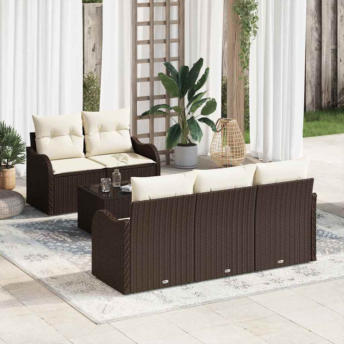 Terrassenset mit braunen Korbmöbeln, cremefarbenen Kissen und einem Couchtisch mit Glasplatte, auf einem Teppich. Pflanzen und Vorhänge im Hintergrund.