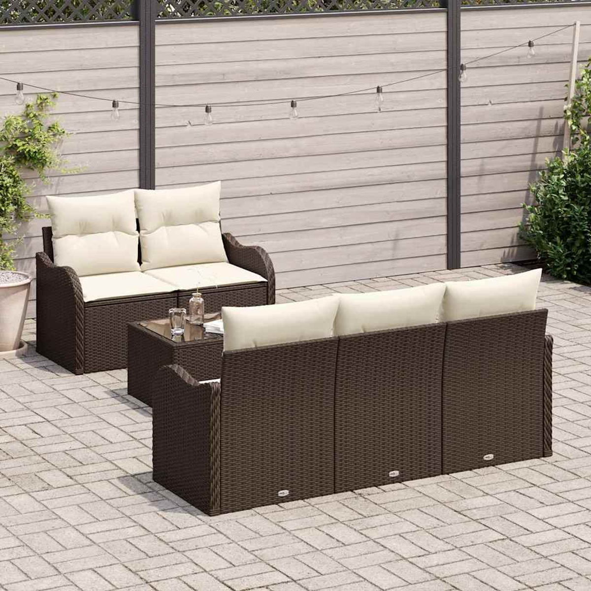Terrassenset: braunes Korbmöbel-Sofa und -Liege mit cremefarbenen Kissen, ein Tisch mit Glasplatte und Lichterketten vor einem Holzzaun.