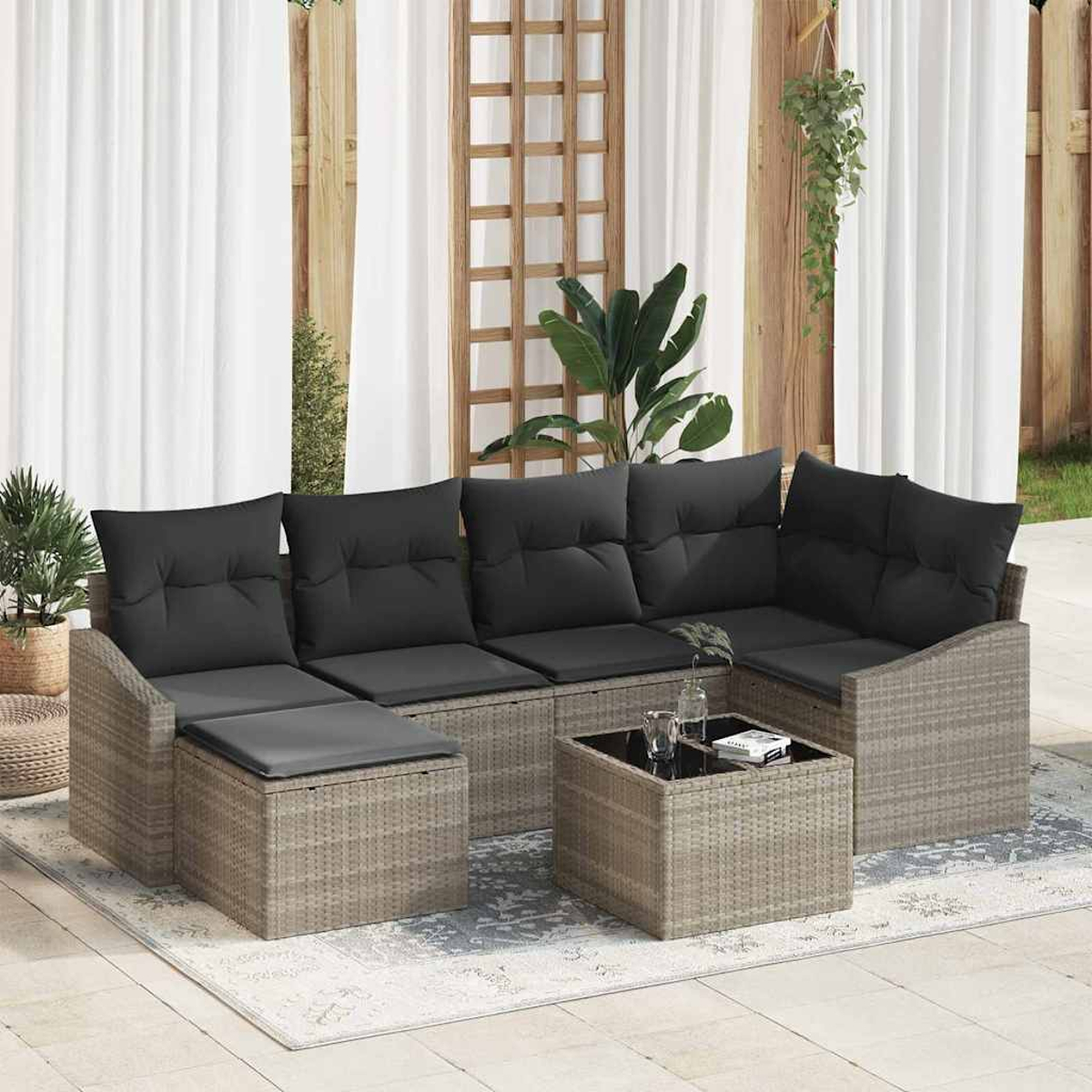 Garten-Rattansofa-Set mit schwarzen Kissen, Couchtisch und Pflanzen. Weiße Vorhänge und Holzgitter im Hintergrund.