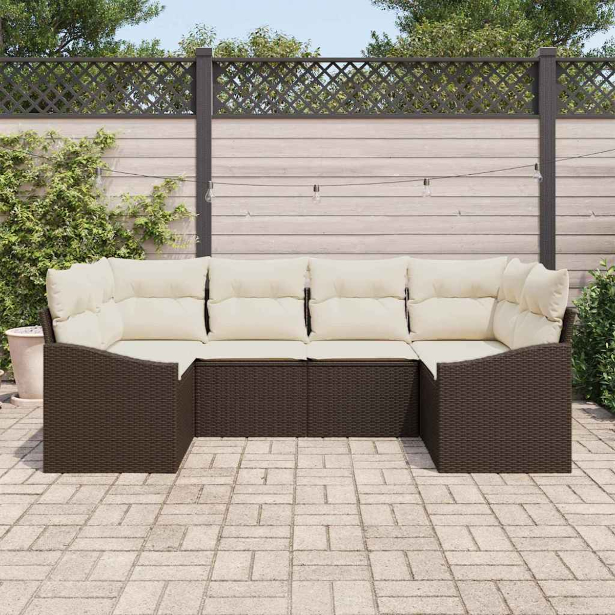 Outdoor-Ecksofa mit cremefarbenen Kissen. Dunkelbraunes Korbgeflecht auf gepflasterter Terrasse, mit Holzzaun und Grün im Hintergrund.