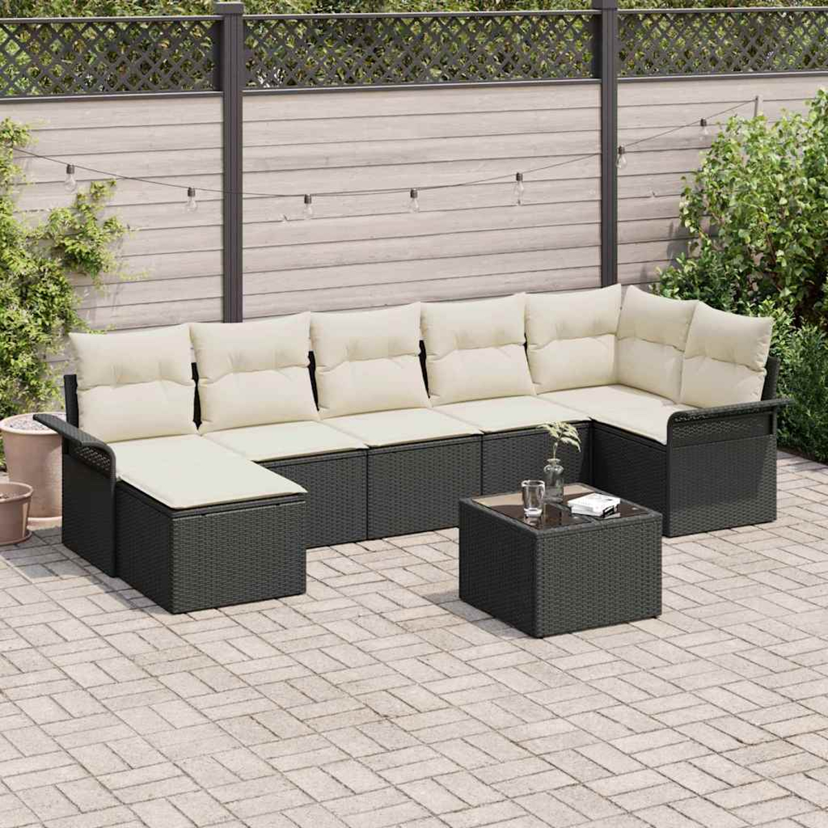 Sofá seccional exterior con cojines crema, estructura de mimbre negro y una mesa cuadrada pequeña.  Sobre un patio de ladrillos con valla de fondo.