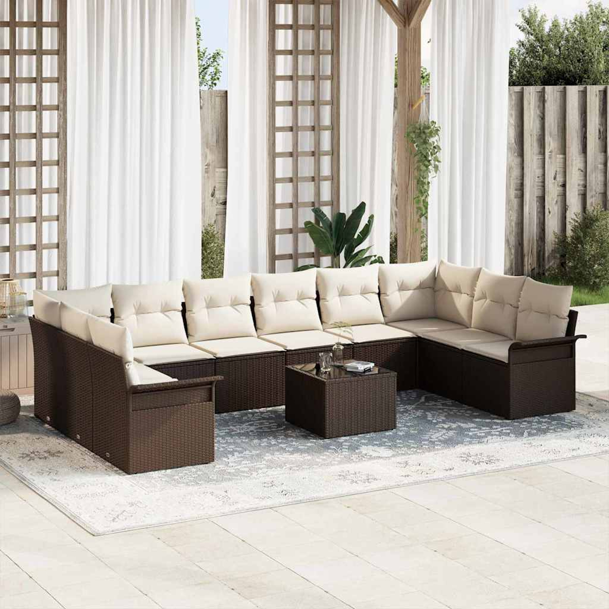 Divano componibile da esterno con cuscini beige, struttura in vimini marrone e tavolino su un tappeto fantasia. Tende bianche sullo sfondo.
