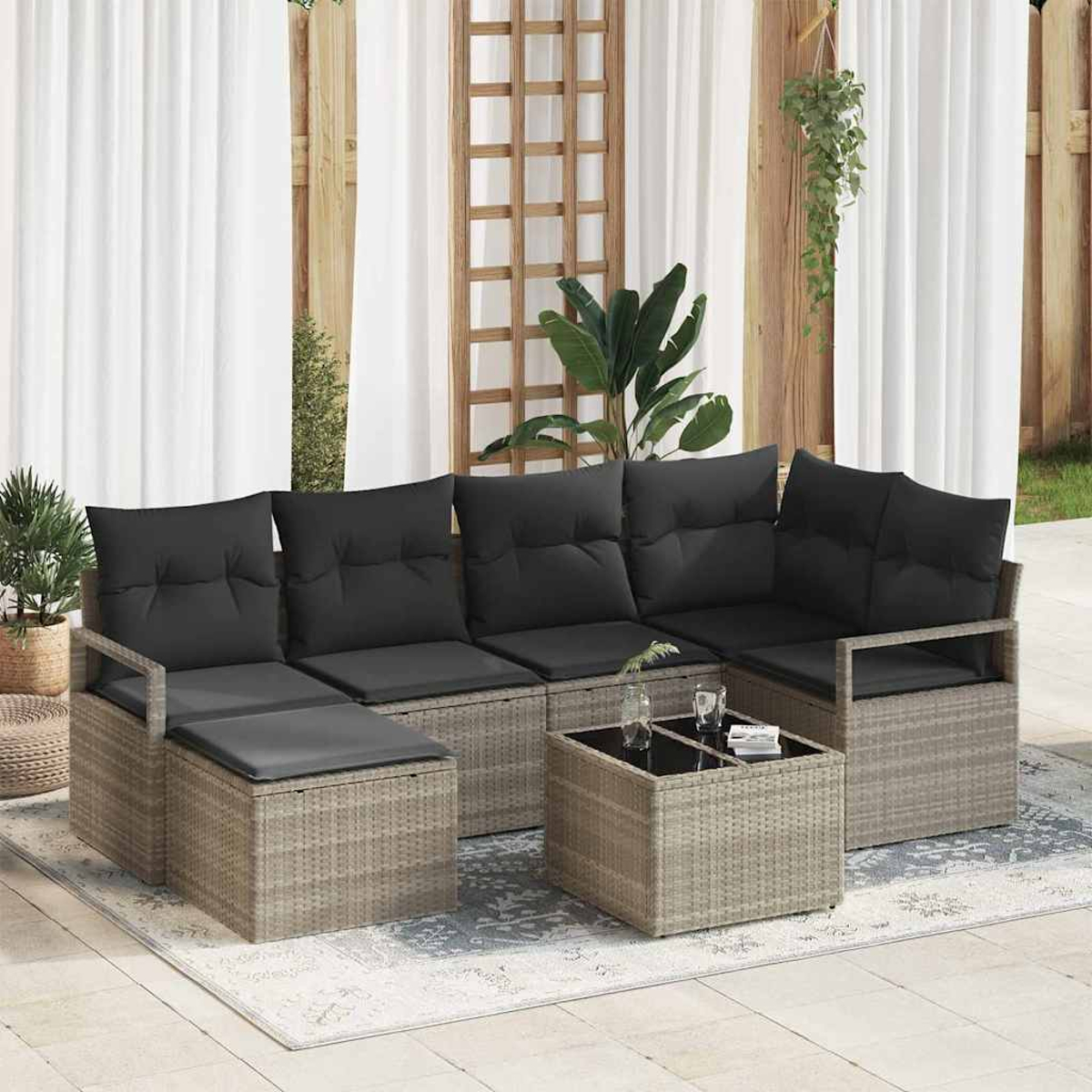 Gartenmöbel-Set aus Rattan mit schwarzen Kissen, einem Couchtisch und Pflanzen. Weiße Vorhänge und Holzgitter im Hintergrund.