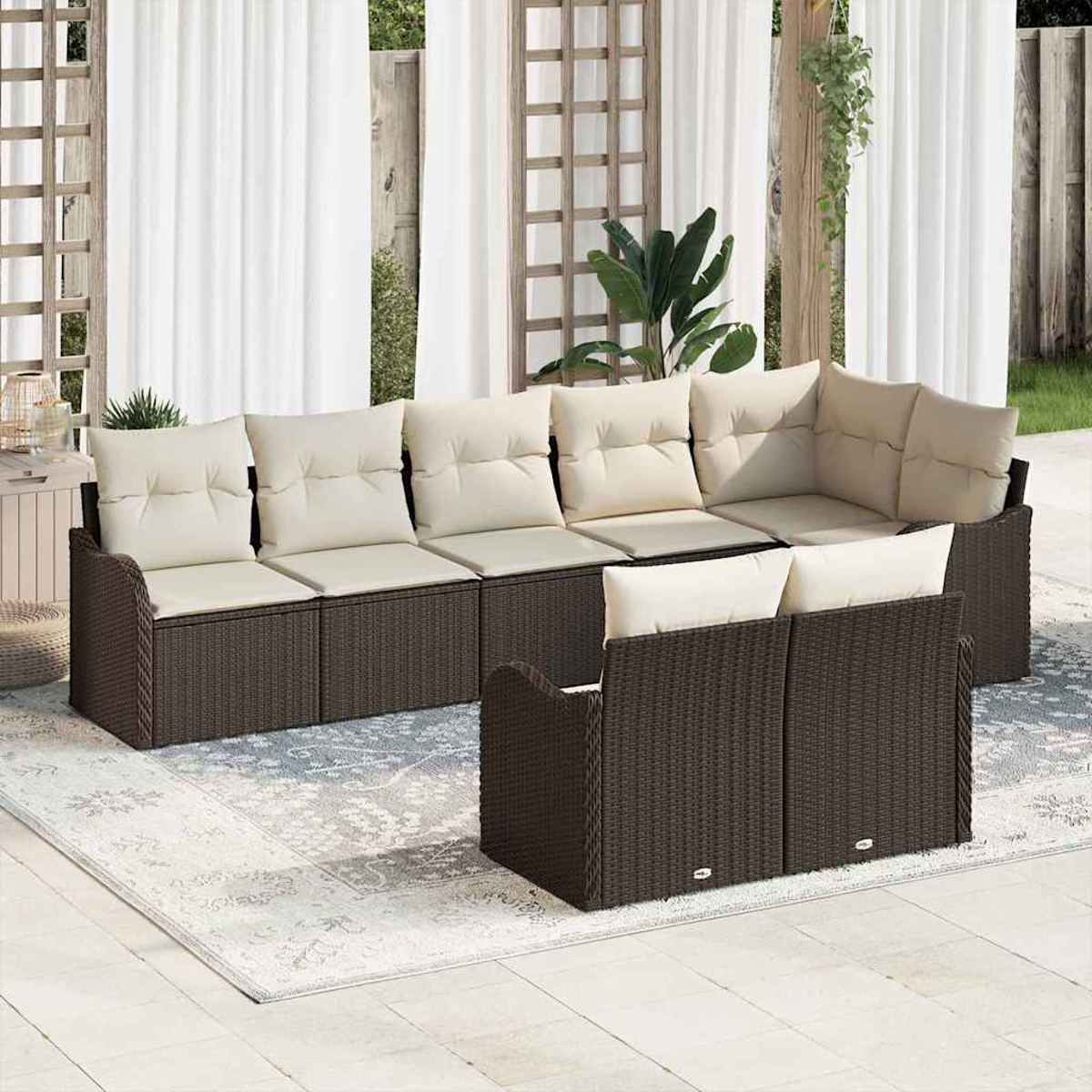 Sofá seccional exterior de mimbre marrón con cojines crema sobre una alfombra estampada. Cortinas blancas y vegetación al fondo.