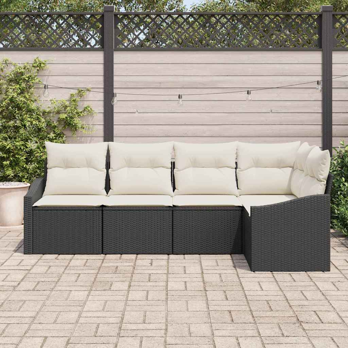 Sofá seccional para exteriores con cojines crema. Estructura de mimbre negro sobre patio de ladrillos, con valla de madera y plantas detrás.