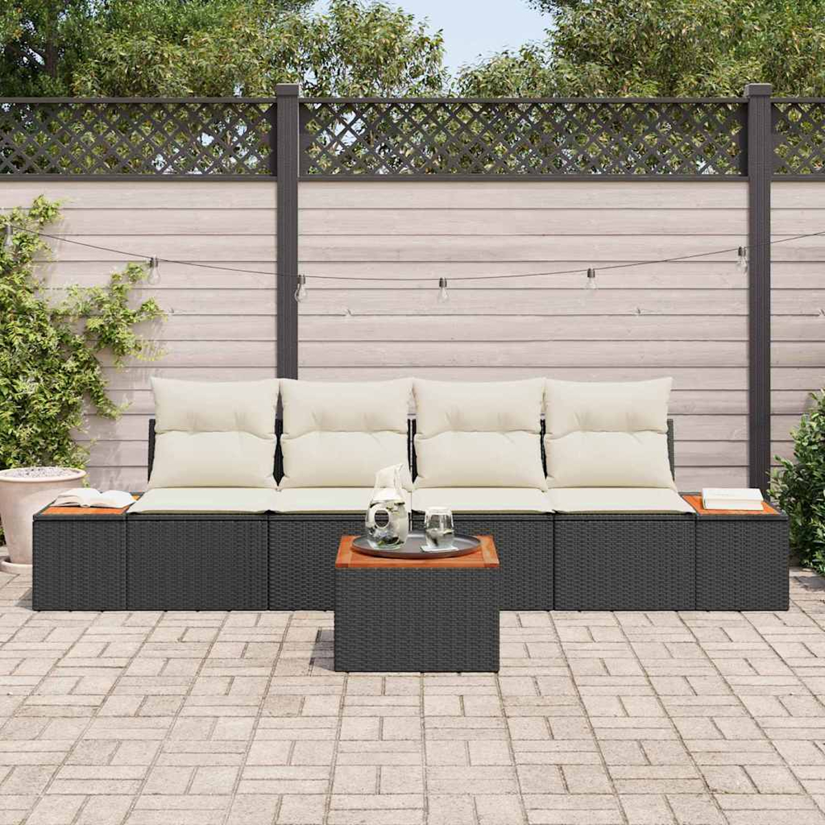 Außenszene mit schwarzem Rattansofa und Tisch, cremefarbenen Kissen, hölzernen Tischplatten und einer grauen Backstein-Terrasse. Im Hintergrund ein Holzzaun.