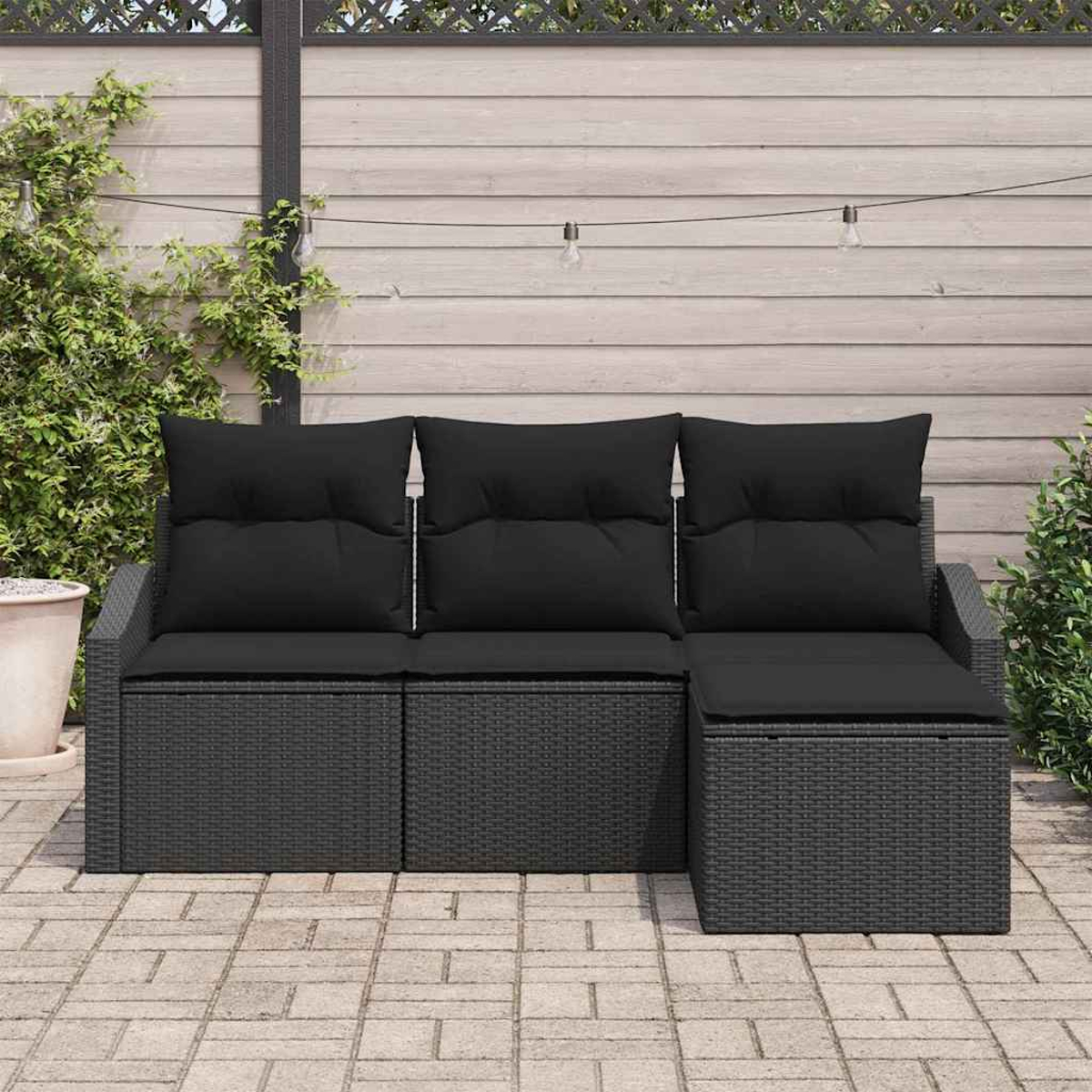 Schwarzes Outdoor-Ecksofa aus Korbgeflecht mit schwarzen Kissen. Auf einer Terrasse mit Holzzaun im Hintergrund. Pflanzen an den Seiten.