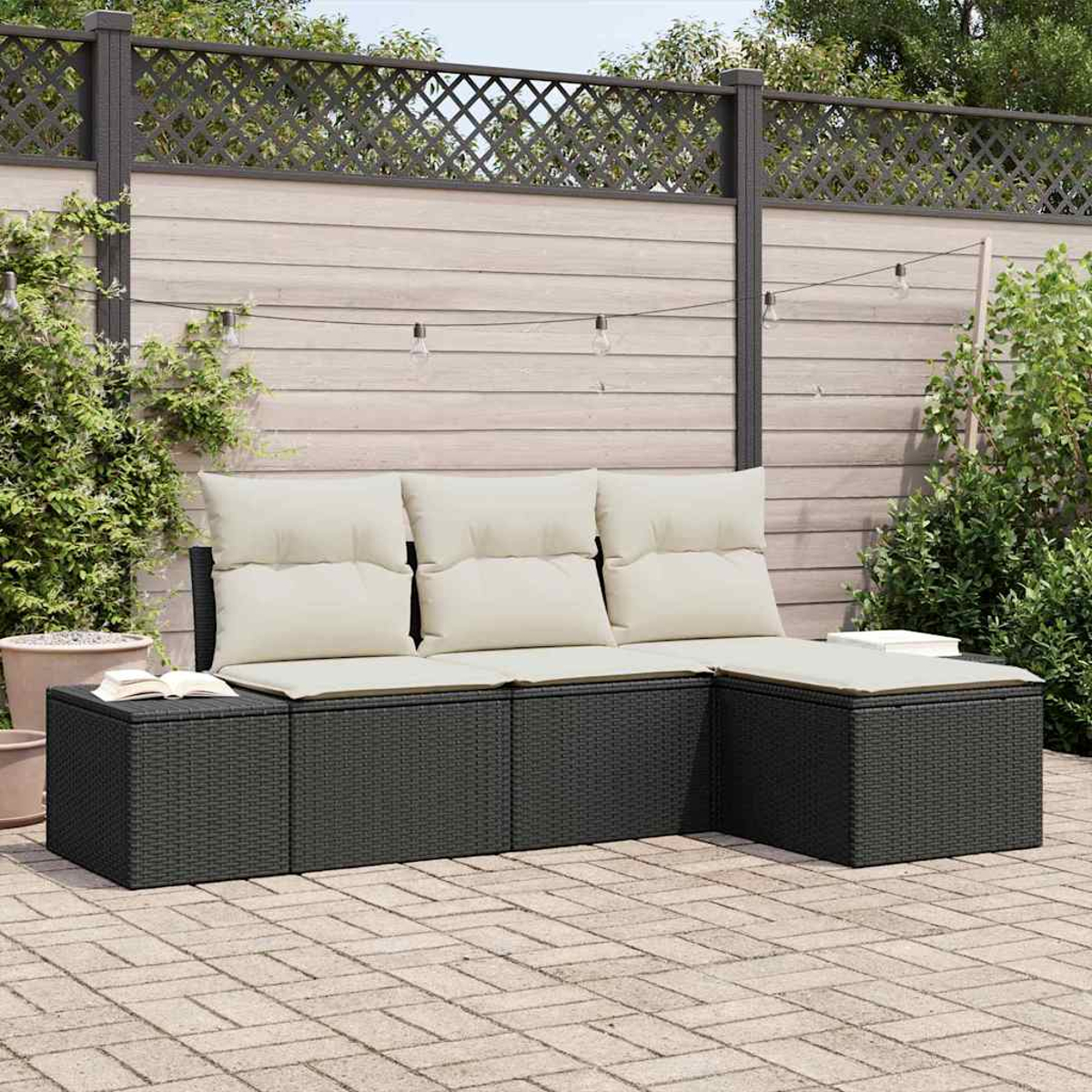 Outdoor-Ecksofa mit cremefarbenen Kissen. Vor einem Holzzaun mit Lichterketten auf einer Backsteinterrasse. Grüne Pflanzen umgeben.