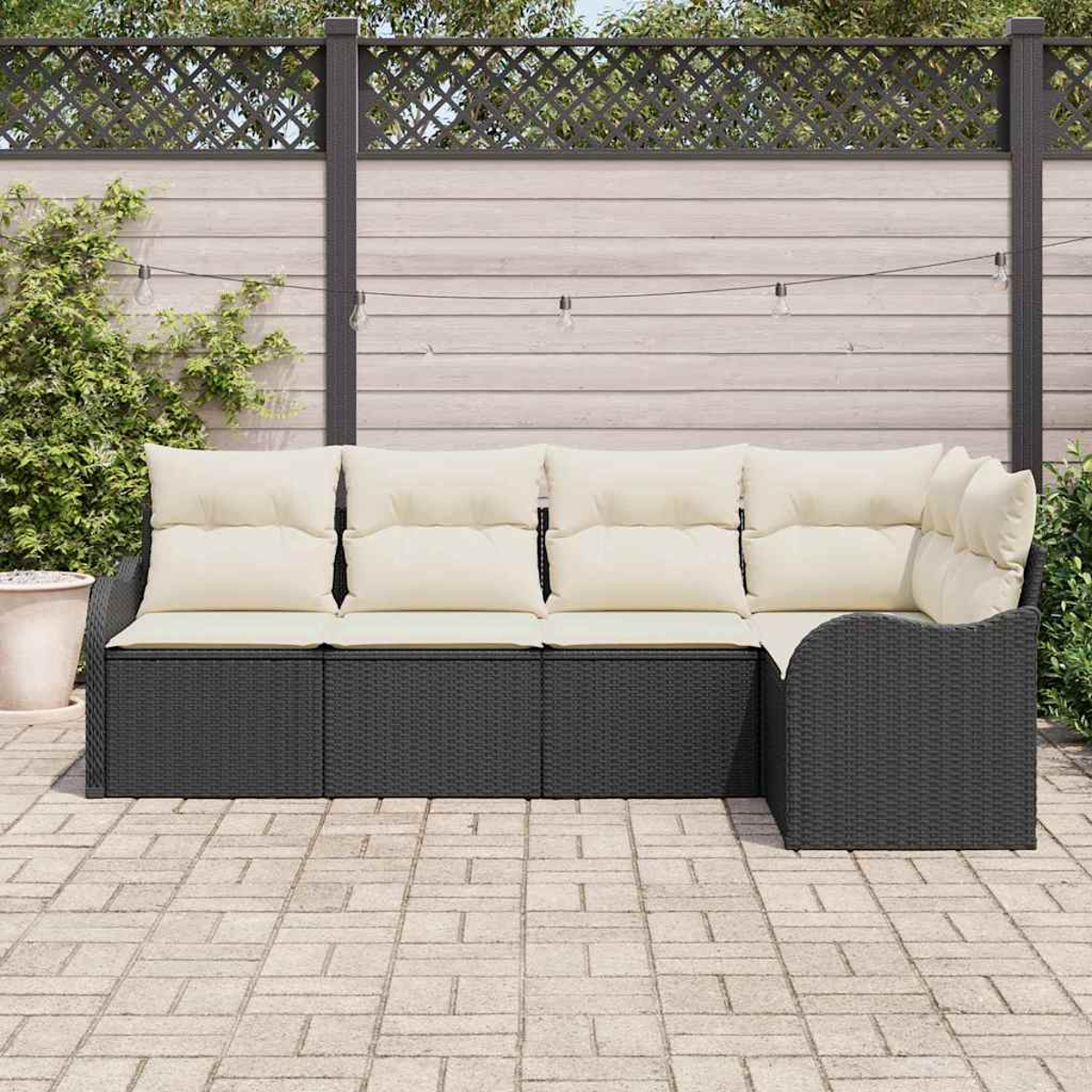 Ein schwarzes Outdoor-Ecksofa aus Korbgeflecht mit cremefarbenen Kissen steht auf einer Backstein-Terrasse vor einem Holzzaun.