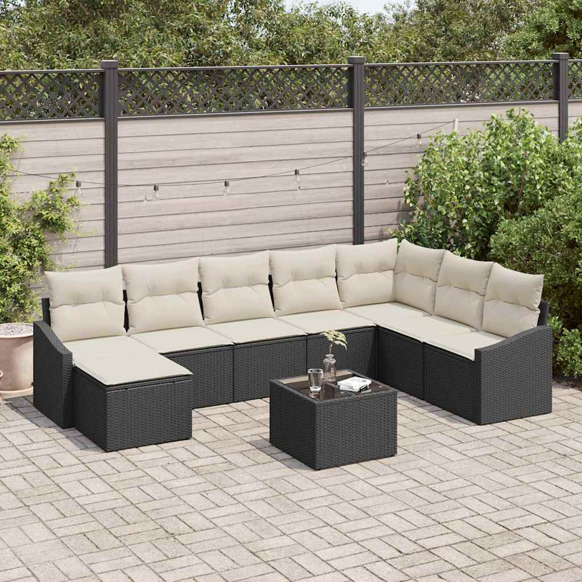 Ein Terrassenset mit dunkelgrauem Korbmöbel und hellbeigen Kissen. Vorne steht ein kleiner Tisch mit Gläsern. Grüne Büsche und ein Zaun dahinter.