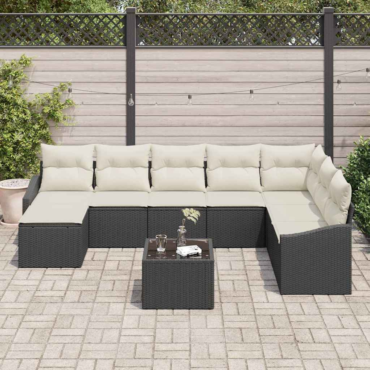 Ein dunkelgraues Rattan-Ecksofa mit cremefarbenen Kissen und einem quadratischen Couchtisch auf einer gepflasterten Terrasse, im Hintergrund ein Zaun.