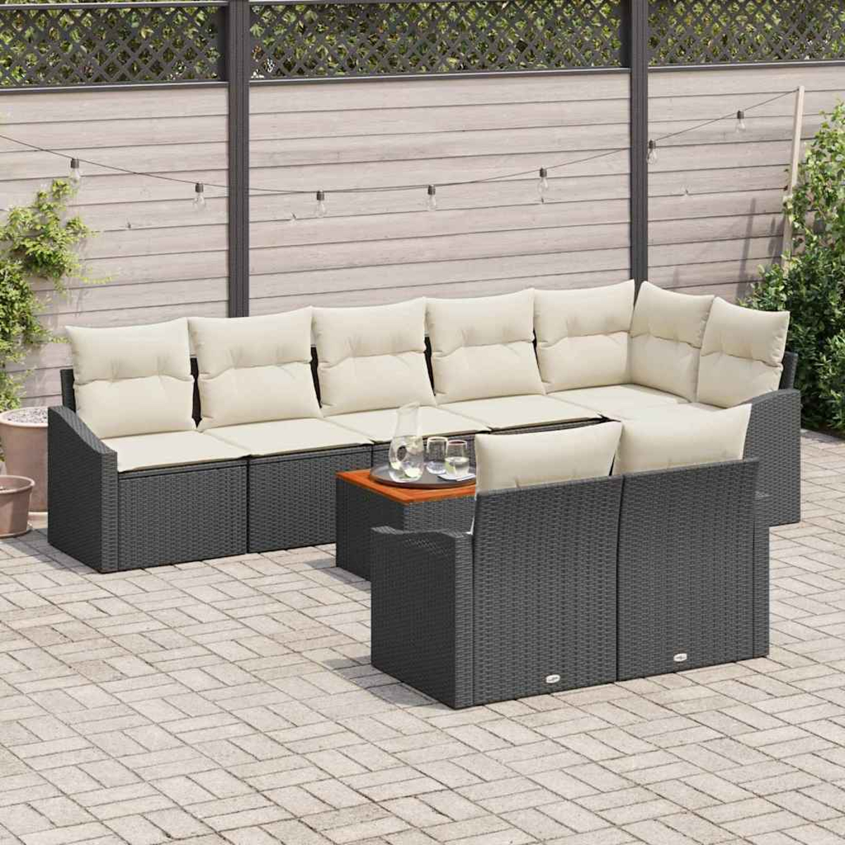 Gartenmöbel-Set. Dunkelgraues Korbmöbel-Sofa mit cremefarbenen Kissen, kleiner Holztisch und Gläser. Hintergrund: Holzzaun.