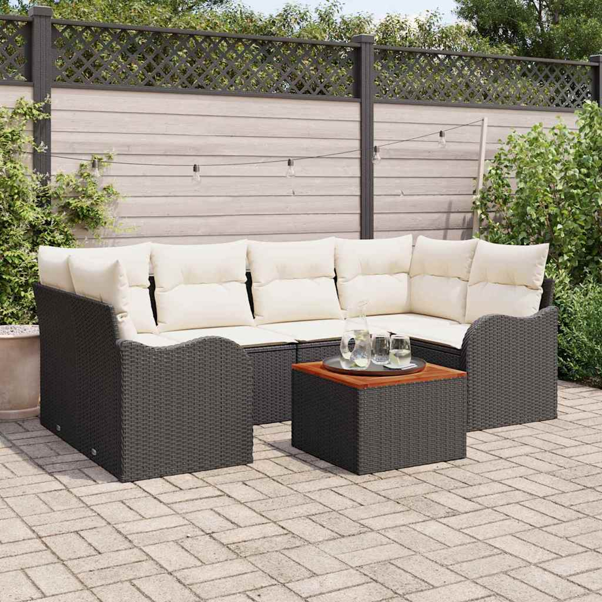 Outdoor-Ecksofa mit cremefarbenen Kissen, ein quadratischer Tisch mit Holzplatte und ein Zaun mit Pflanzen und Lichtern im Hintergrund.