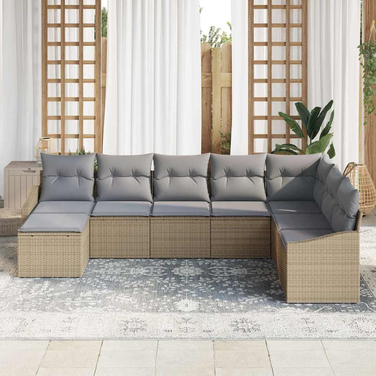 Ein beiges Ecksofa aus Korbgeflecht mit grauen Kissen steht auf einem gemusterten Teppich. Dahinter sind ein Holzgitter, weiße Vorhänge und Pflanzen.