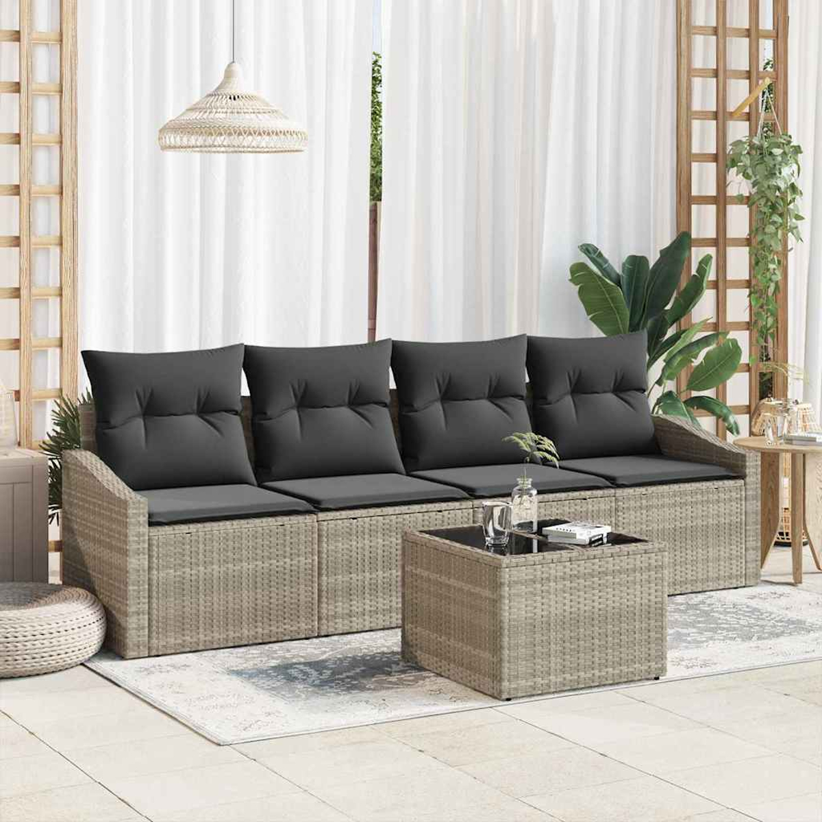 Garten-Korbsofa mit dunklen Kissen, Glastisch und weißen Vorhängen. Pflanzen und Holzgitter im Hintergrund.