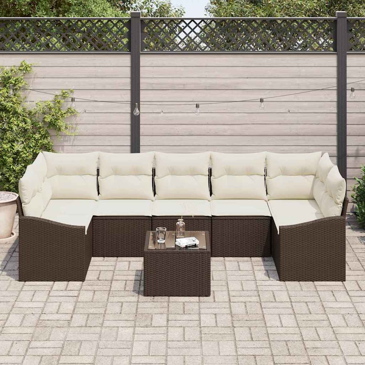 Garten-Sitzgruppe mit cremefarbenen Kissen und braunem Korbgewebe auf einer gepflasterten Terrasse. Davor steht ein kleiner Tisch mit Glasplatte.