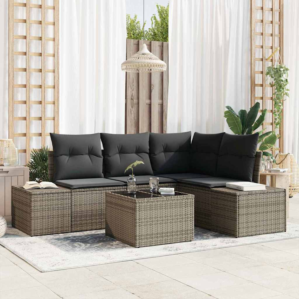 Wicker-Sofa mit schwarzen Kissen und einem Tisch mit Glasplatte auf einer Terrasse. Weiße Vorhänge und grüne Pflanzen im Hintergrund.