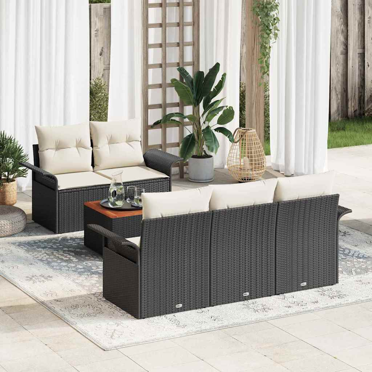 Garten-Terrassenset mit schwarzen Korbmöbeln, cremefarbenen Kissen und einem Holztisch. Ein Teppich und Pflanzen runden die Szene ab.