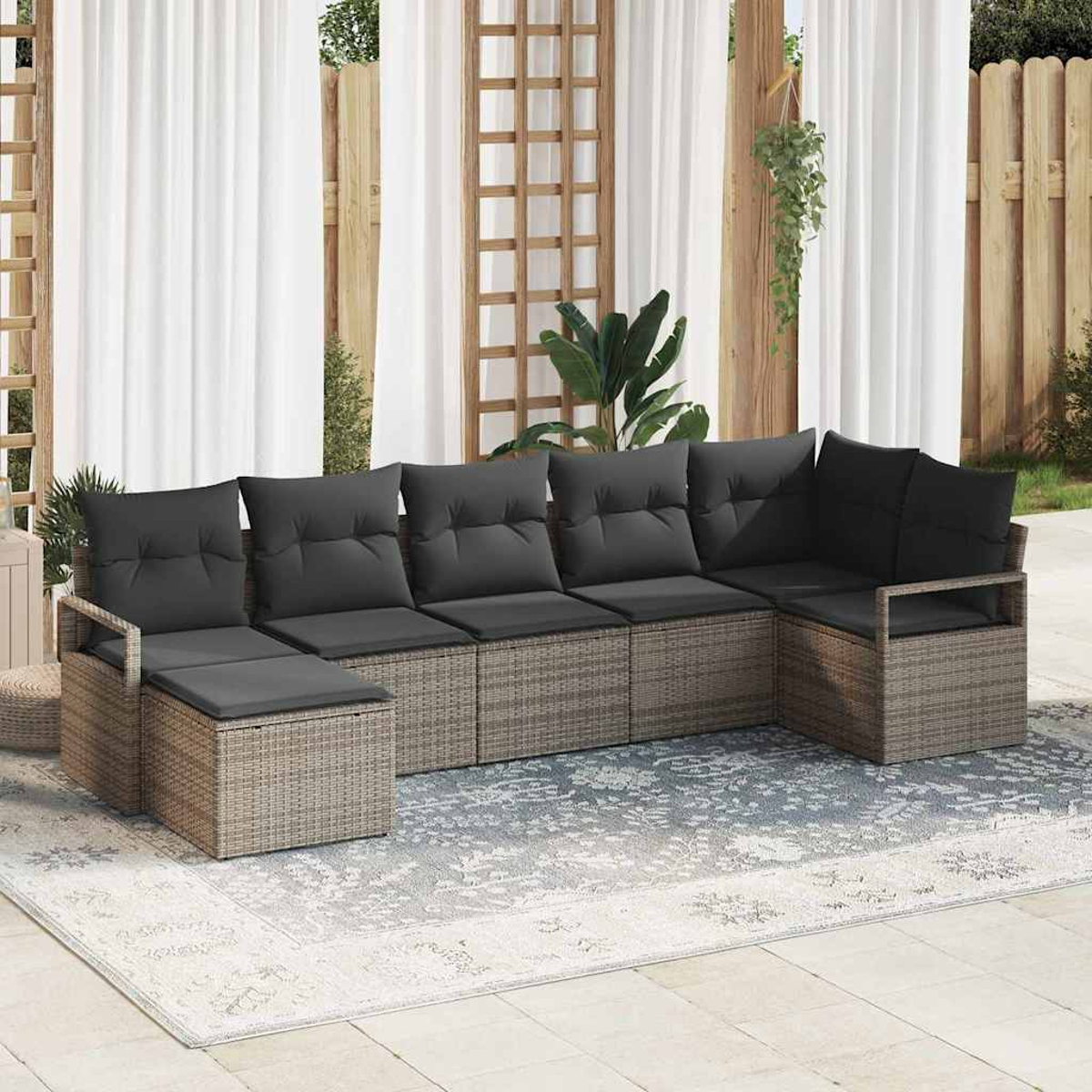 Ein Ecksofa im Freien mit grauen Kissen und einem korbähnlichen Rahmen auf einem gemusterten Teppich. Weiße Vorhänge und ein Holzzaun im Hintergrund.