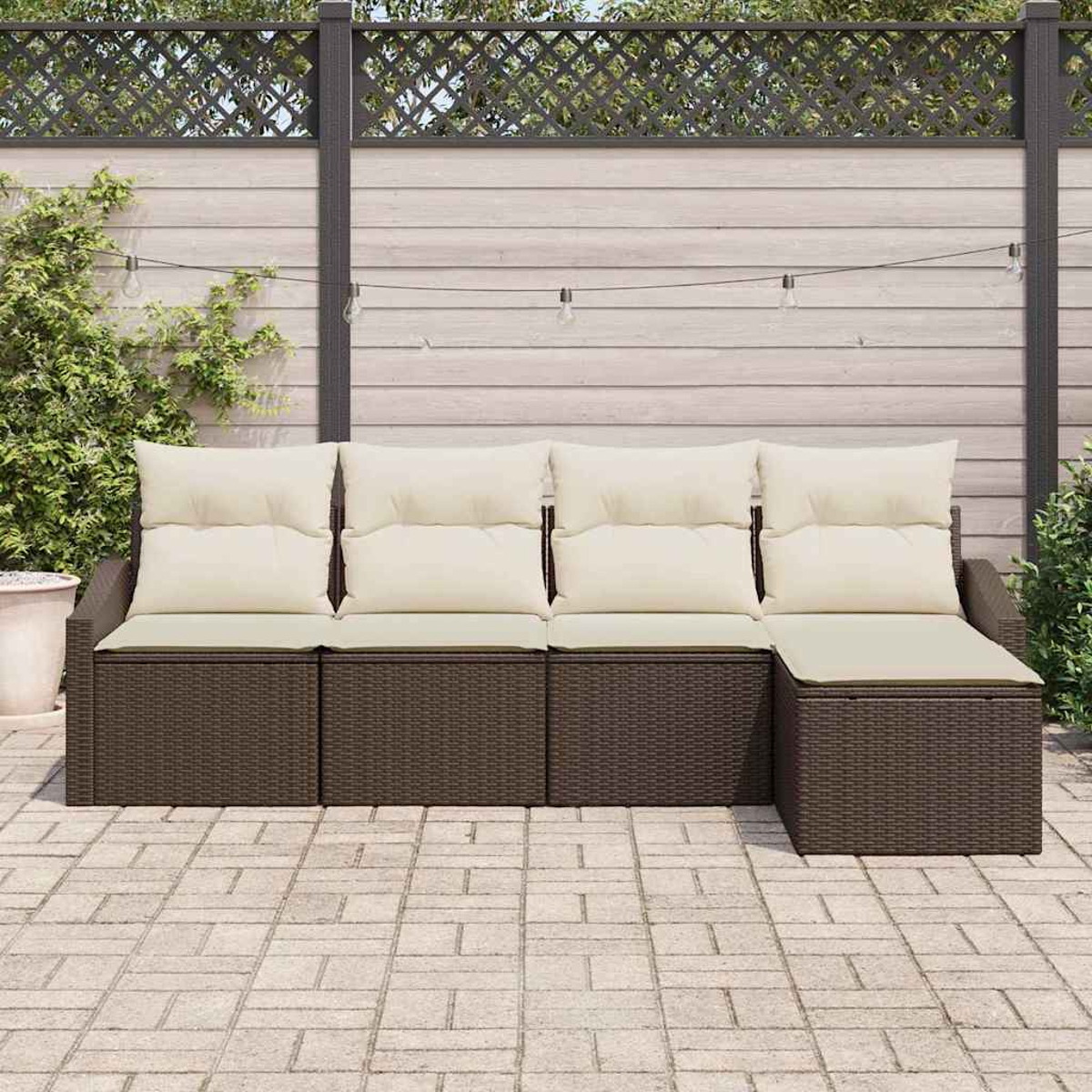 Braunes Rattan-Ecksofa im Freien mit cremefarbenen Kissen auf einer Steinterrasse. Im Hintergrund ein Holzzaun und Grün.