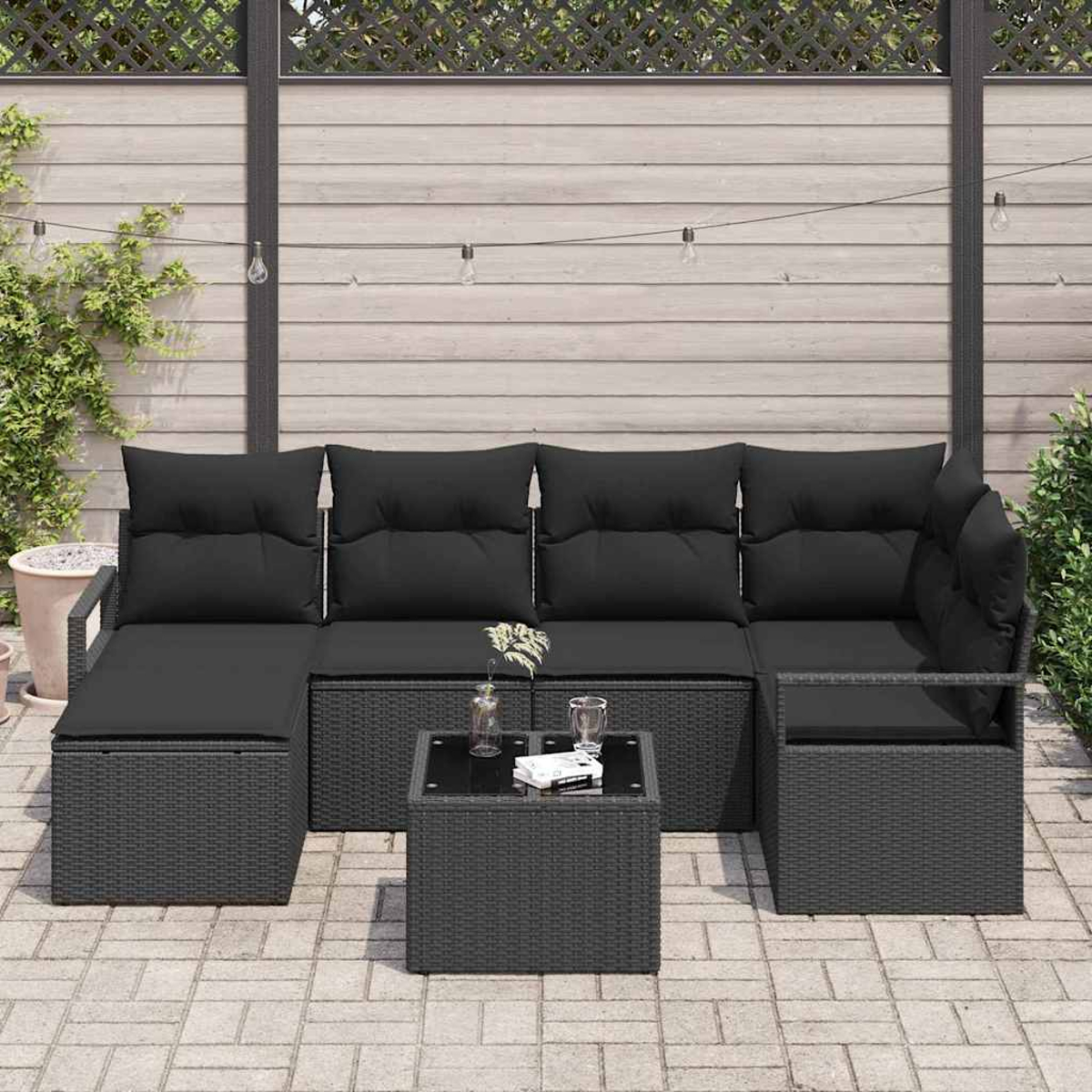 Ein schwarzes Outdoor-Sofa aus Korbgeflecht mit Kissen, ein quadratischer Tisch und ein Holzzaun im Hintergrund, mit dekorativen Lichtern.