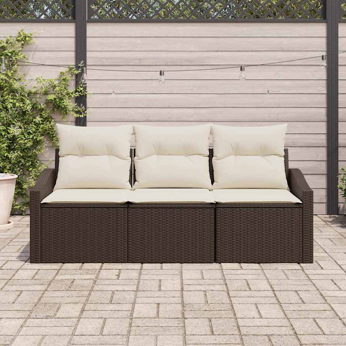 Braunes Rattan-Sofa mit cremefarbenen Kissen auf einer Steinterrasse. Im Hintergrund ein Holzzaun und Lichterketten mit grünen Pflanzen.