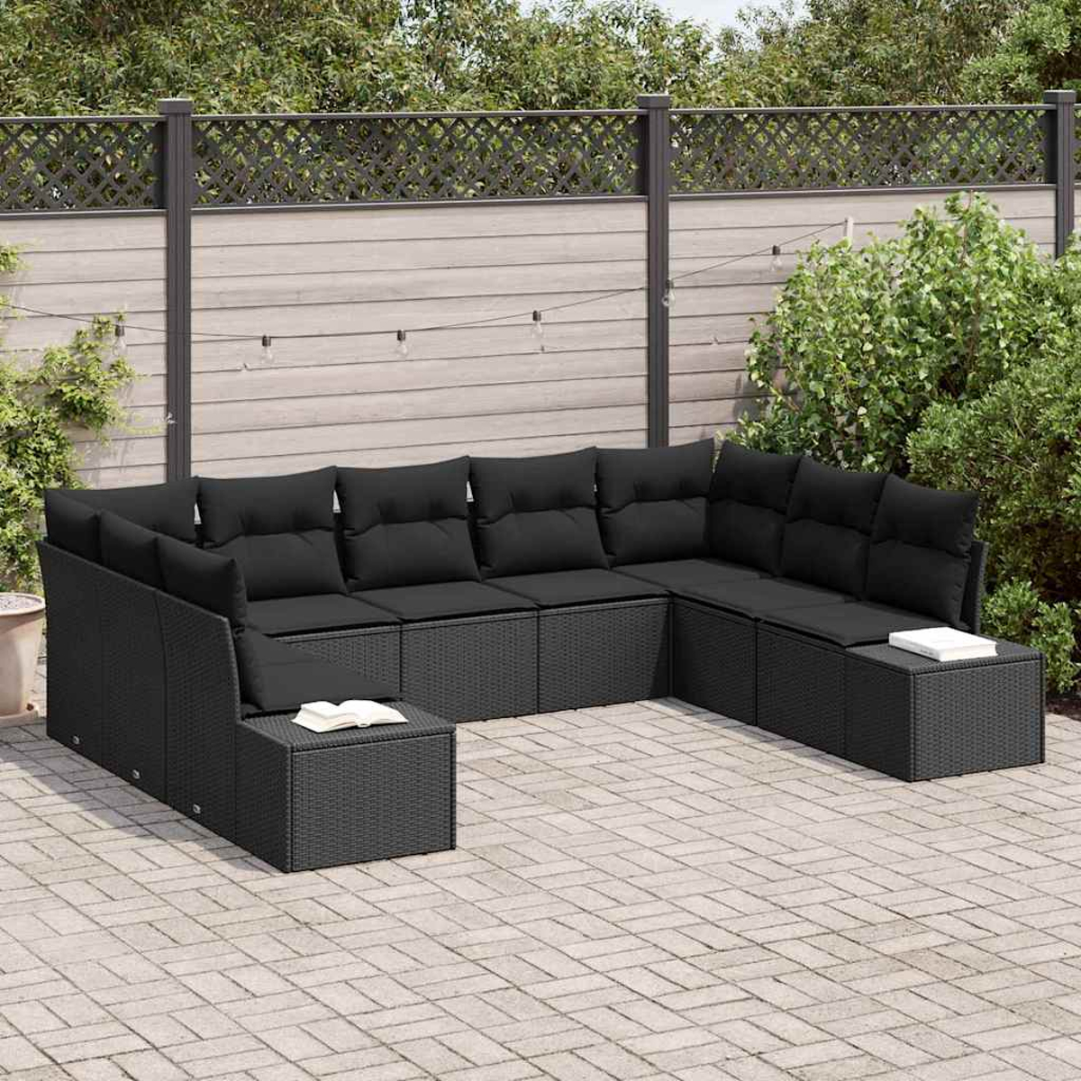 Ein schwarzes Ecksofa aus Korbgeflecht mit schwarzen Kissen steht auf einer Ziegelsteinterrasse. Ein Holzzaun und Grün bilden den Hintergrund.