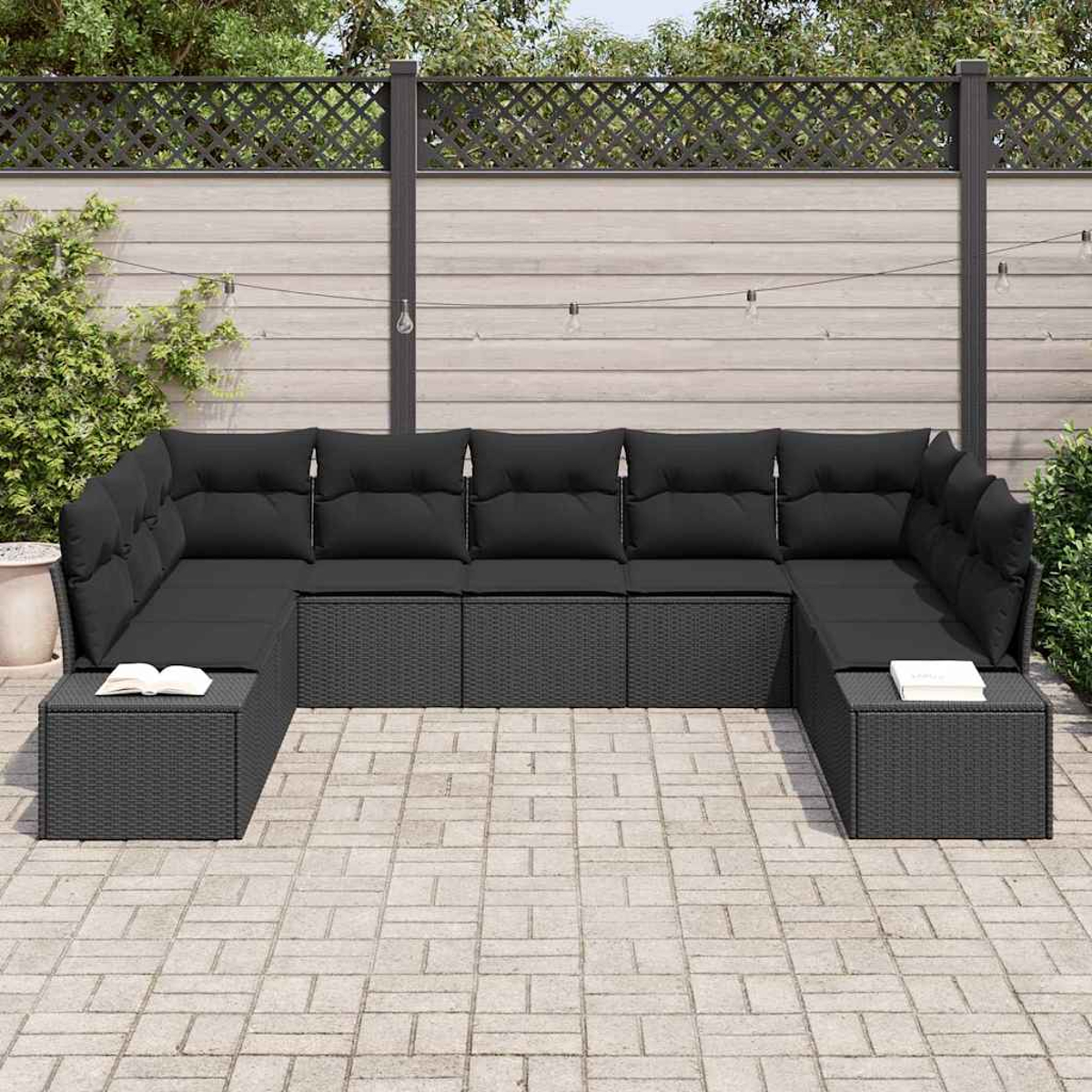 Ein schwarzes Korbsessel-Sofa mit schwarzen Kissen steht auf einer Terrasse. Auf jedem Ende liegt ein Buch. Im Hintergrund sind ein Zaun und Pflanzen.