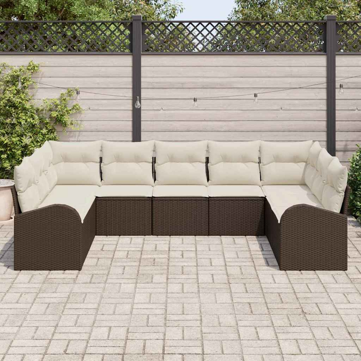 Ein braunes Ecksofa aus Korbgeflecht mit cremefarbenen Kissen steht auf einer Terrasse. Im Hintergrund sind ein Holzzaun und Grün zu sehen.