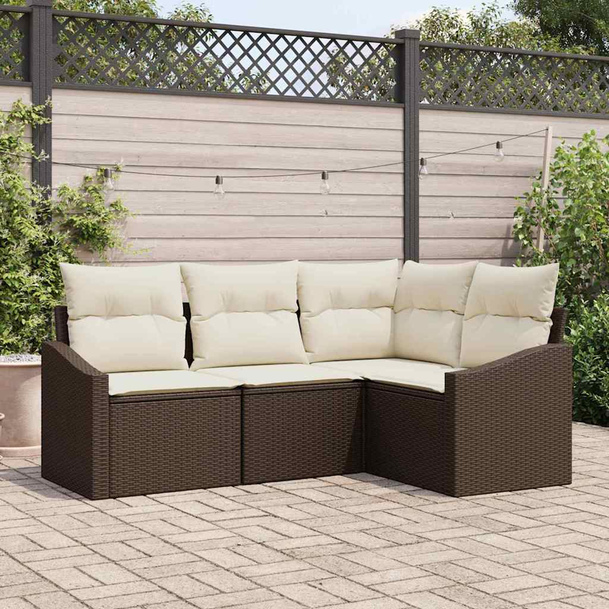 Ein braunes Korbsessel-Ecksofa mit cremefarbenen Kissen steht auf einer Ziegelsteinterrasse. Ein Holzzaun mit Lichterketten ist im Hintergrund.