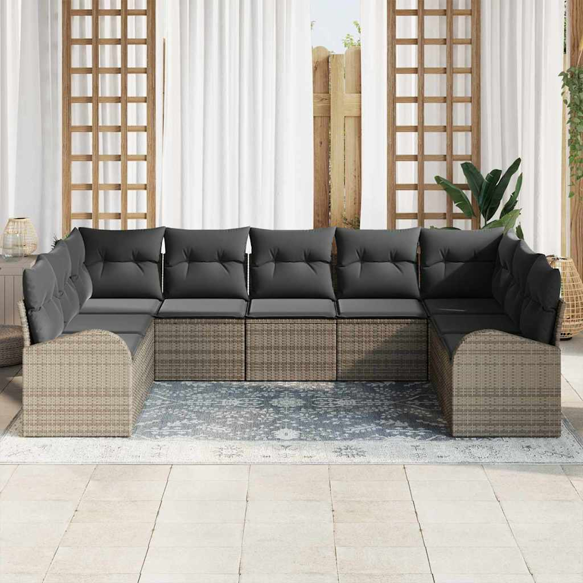 Ein geflochtenes Outdoor-Ecksofa mit dunkelgrauen Kissen steht auf einem gemusterten Teppich. Weiße Vorhänge und Holzgitter im Hintergrund.
