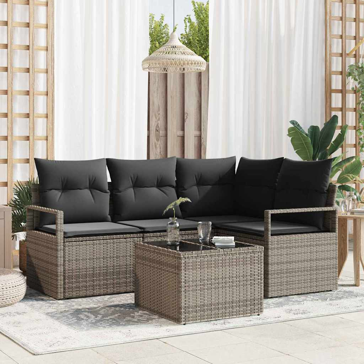 Rattansofa im Freien mit schwarzen Kissen und einem Glastisch. Weiße Vorhänge, eine Hängelampe und Pflanzen im Hintergrund.